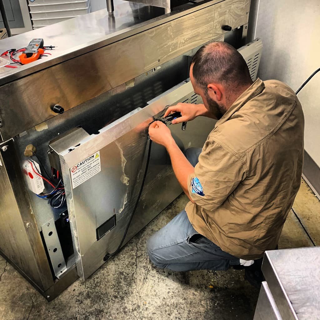 Technician repairing a commercial appliance in a professional kitchen environment, focusing on wiring and using tools for precise adjustments