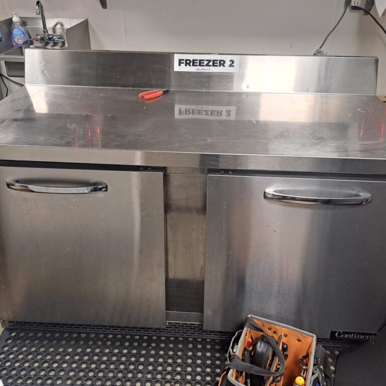 A stainless steel commercial freezer with two doors, labeled "Freezer 2," located in a kitchen environment. A red-handled tool is placed on top of the freezer, and a tool bag is visible in the lower-right corner.