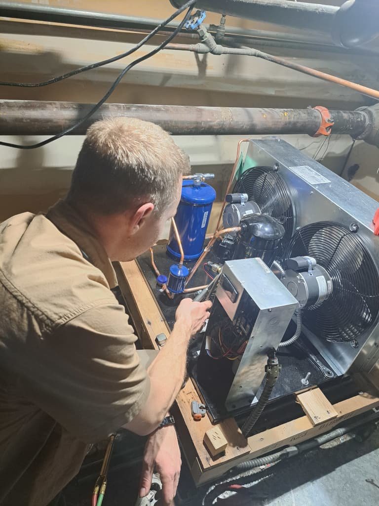 A technician in a khaki shirt performing diagnostics on an HVAC system, examining electrical components with surrounding pipes and system parts in a mechanical room