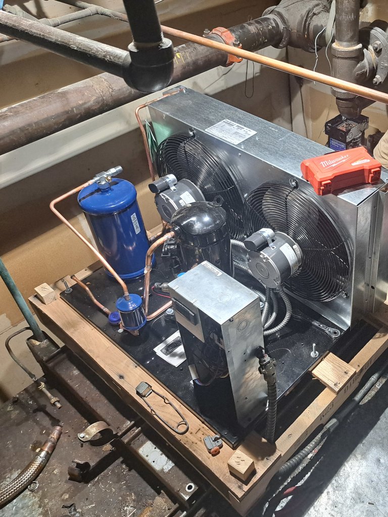 An HVAC unit with dual fans and compressor components mounted on a wooden base, surrounded by copper piping and industrial plumbing in a mechanical room, with a Milwaukee tool case on top