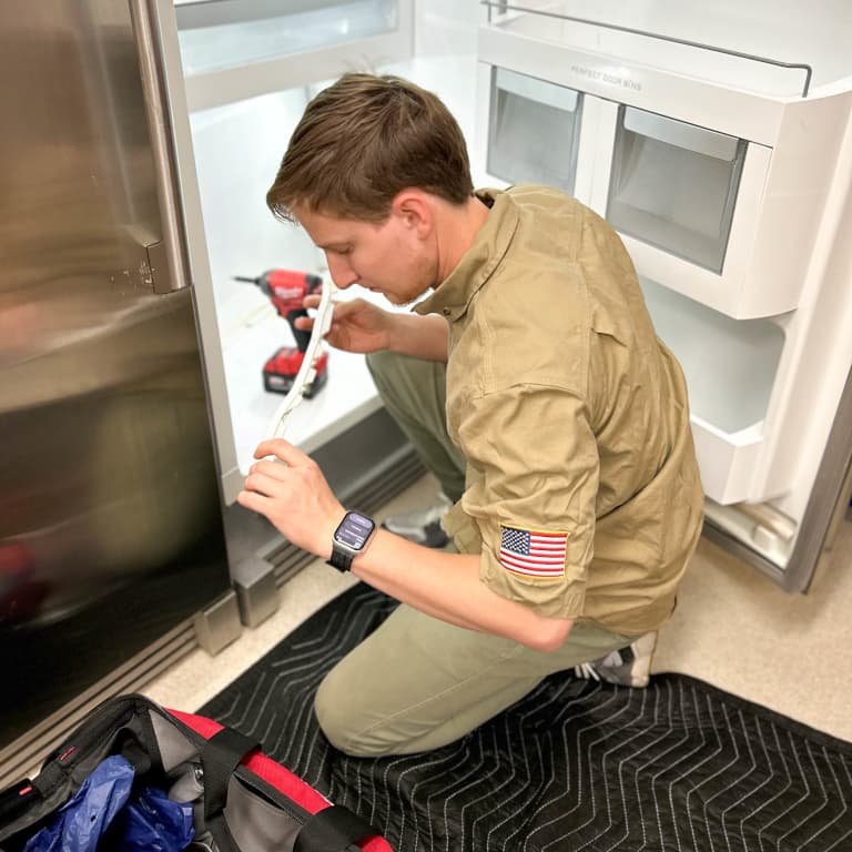 Technician repairing a commercial refrigerator with tools in hand, wearing a khaki uniform with an American flag patch, kneeling on a protective mat, and inspecting refrigerator components