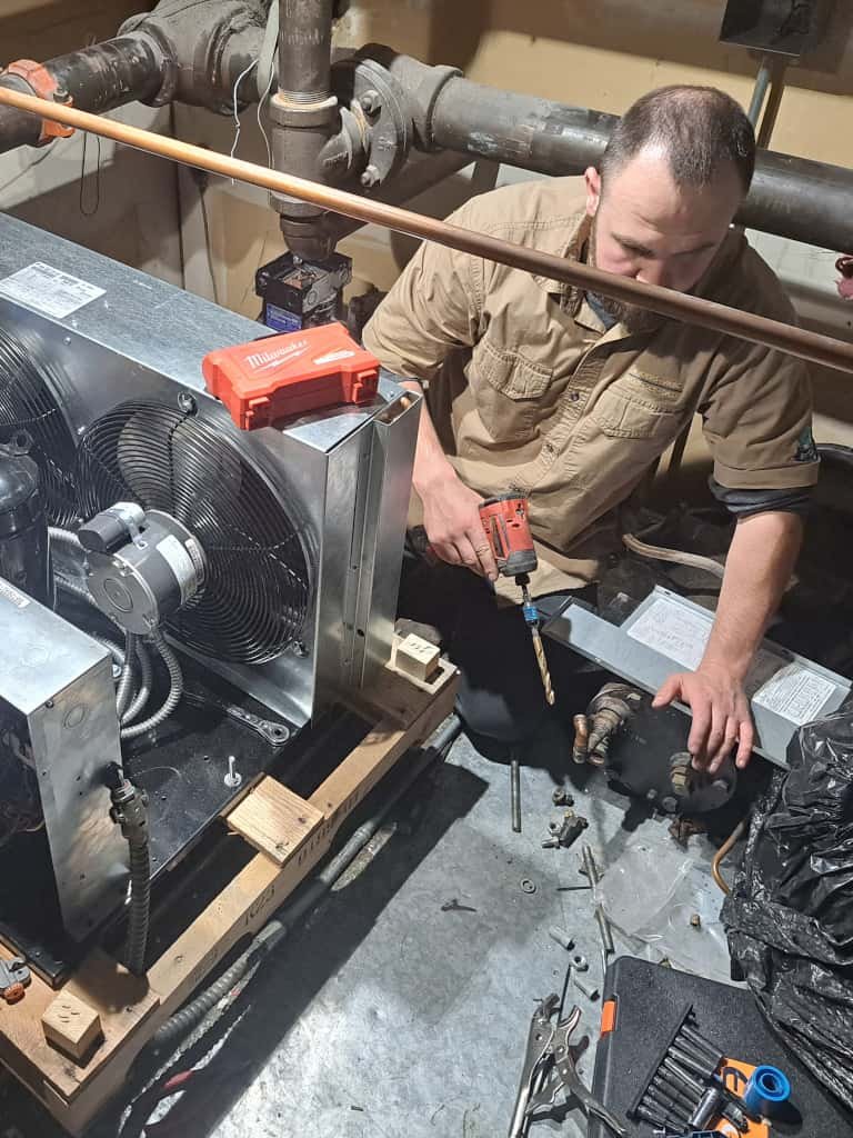 A technician in a khaki shirt working on an HVAC system in a mechanical room, using a Milwaukee drill and surrounded by tools, pipes, and system components.