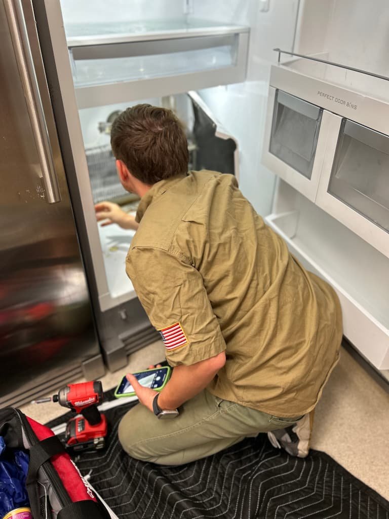 A technician in a khaki shirt with a U.S. flag patch working on a refrigerator, using a Milwaukee screwdriver and diagnostic device, with tools nearby on a protective mat